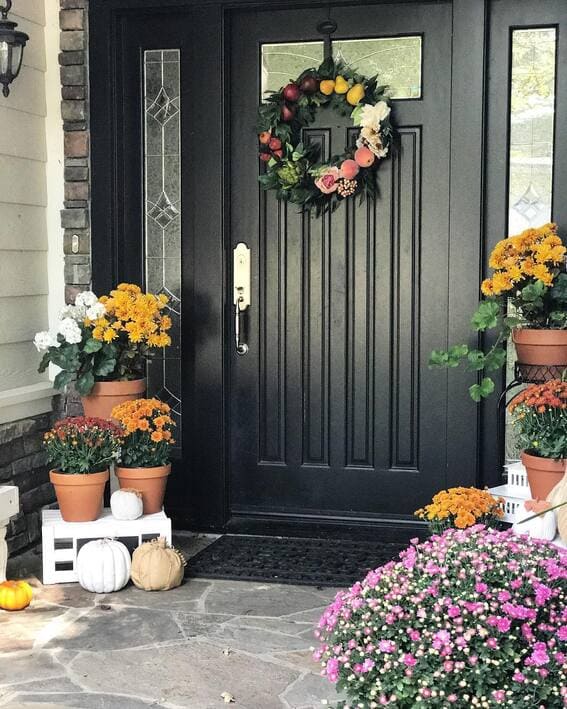 Black front door with a floral wreath, potted chrysanthemums, and white pumpkins decorating the porch.