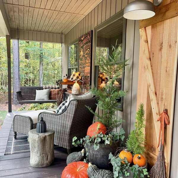Wood-paneled siding, a wicker chair, plaid pillows, rustic Halloween décor and potted evergreens with pumpkins on a forest-side porch