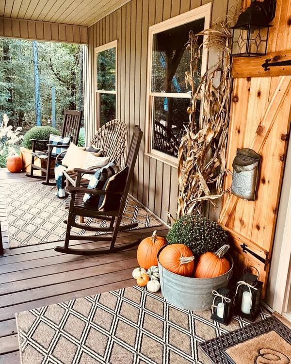 A rustic porch outfitted with rocking chairs, layered rugs, plaid pillows, corn stalks and a galvanized tub filled with pumpkins and mums.