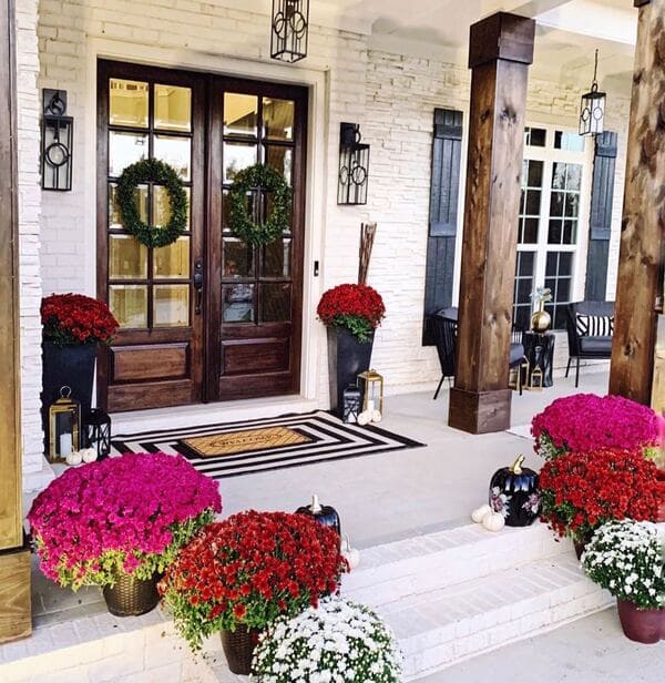 A brick front porch with rich wood doors, fall mums in vibrant kinds, tall black planters, and tiered lanterns.