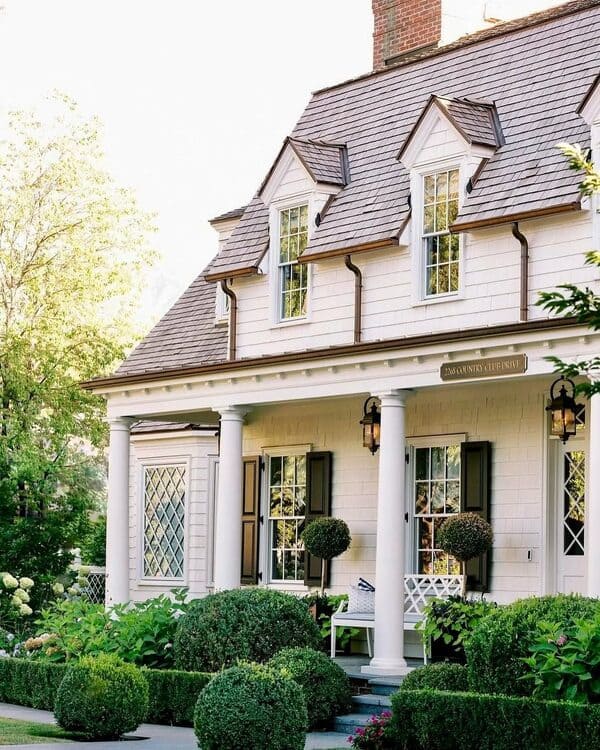 A classic colonial-style house featuring white columns, diamond-pane windows, black shutters, and sculpted greenery on a traditional front porch.