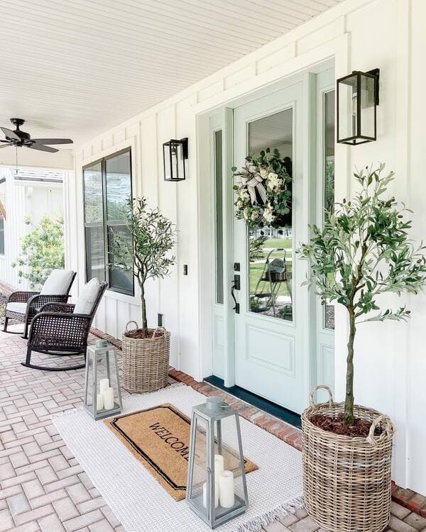 A farmhouse-style porch with board-and-batten siding, mint green front doors, wicker planters holding olive trees and oversized candle lanterns.