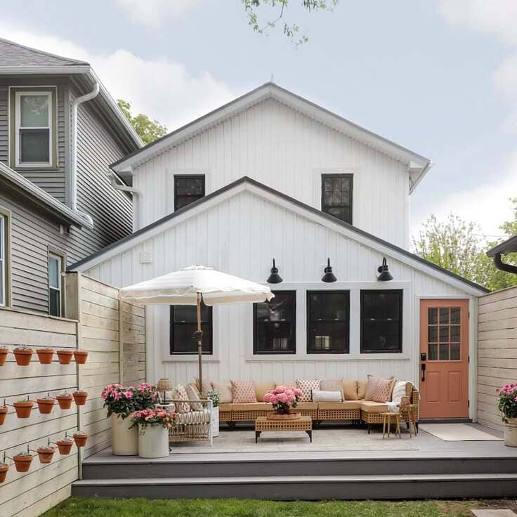 A cozy patio with a white house exterior, wicker furniture, a pink door, and pink flowers on a coffee table.