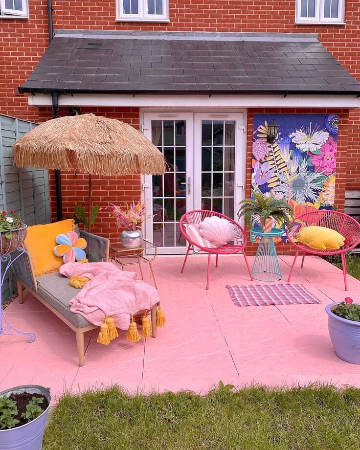 A lively pink patio featuring bamboo chairs, a thatched umbrella, and a bright floral mural against a red brick house.
