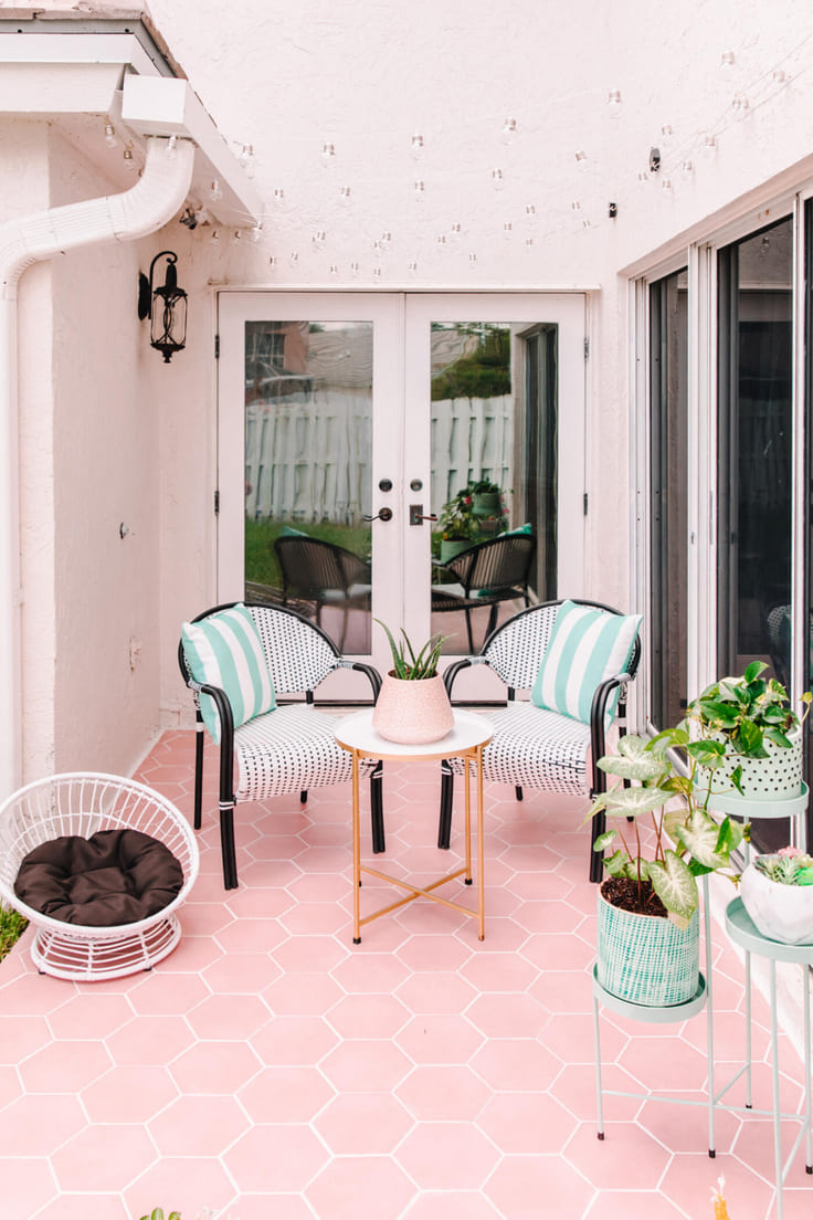 A cozy outdoor patio with pink hexagonal tiles, mint green chairs with striped pillows, a small white and gold table, and potted greenery.