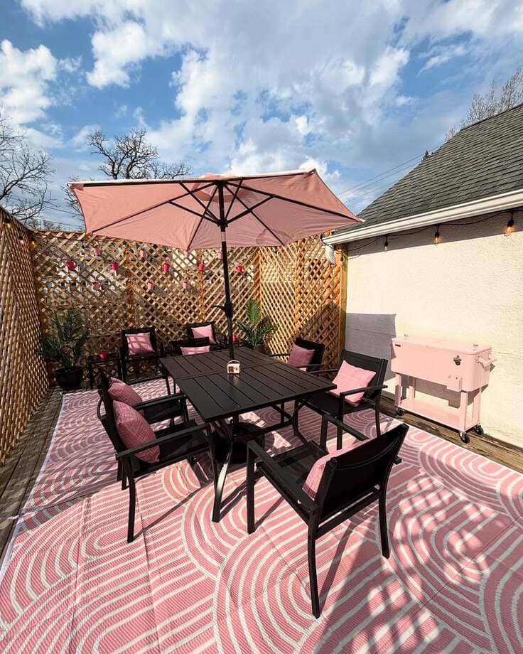 A modern outdoor patio featuring a pink and white patterned rug, black dining set, pink pillows, pink umbrella, and a pink cooler.