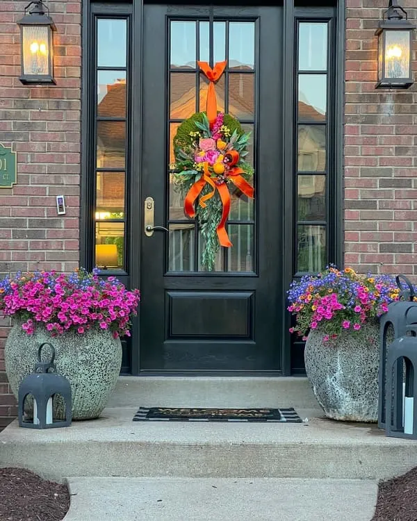 Brick facade dotted with a black door, bright floral wreath and colorful planters.