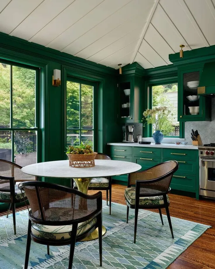 Bold kitchen with rich green cabinetry, floor-to-ceiling windows, white marble countertops, and cane dining chairs