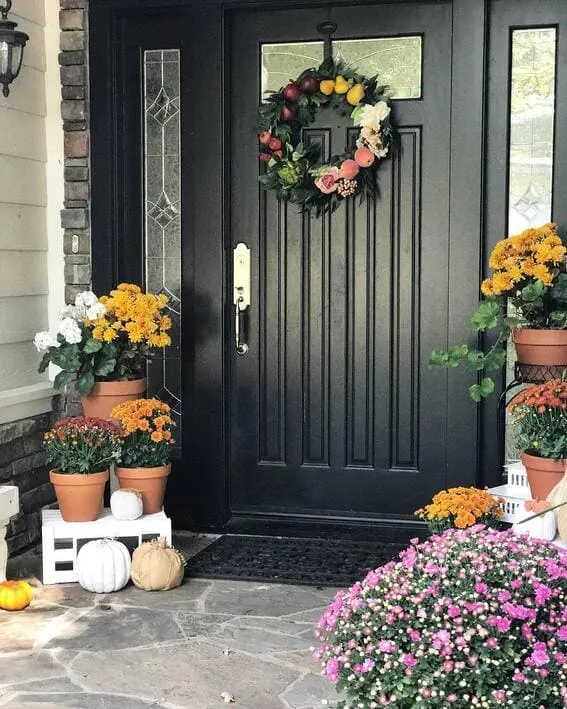 Black front door with a floral wreath, potted chrysanthemums, and white pumpkins decorating the porch.