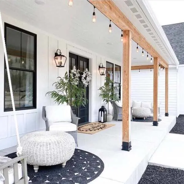 A white farmhouse-style front porch with black doors, wooden beams, wicker furniture, and string lights.