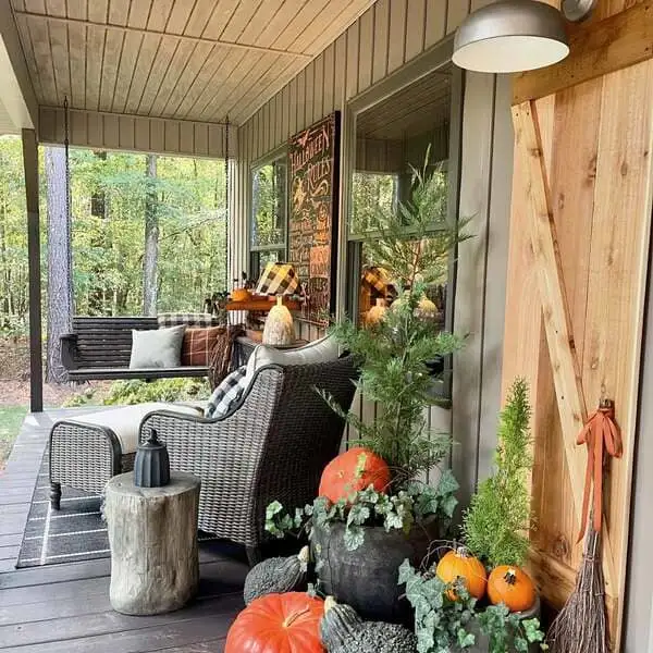 Wood-paneled siding, a wicker chair, plaid pillows, rustic Halloween décor and potted evergreens with pumpkins on a forest-side porch