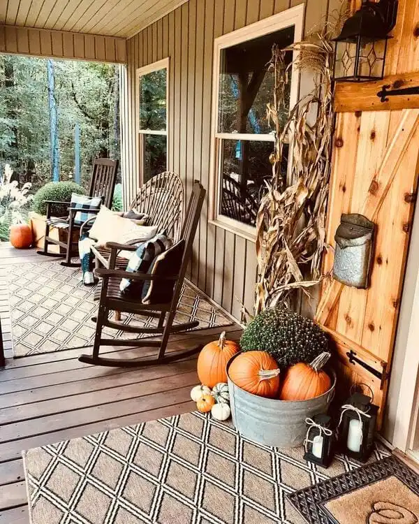 A rustic porch outfitted with rocking chairs, layered rugs, plaid pillows, corn stalks and a galvanized tub filled with pumpkins and mums.