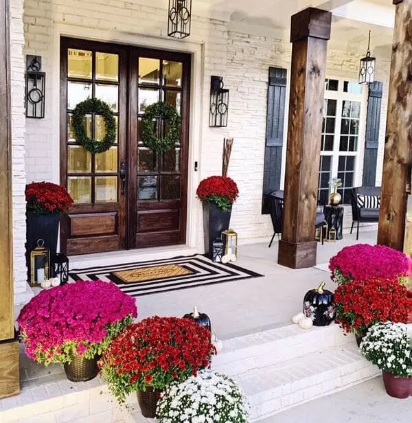 A brick front porch with rich wood doors, fall mums in vibrant kinds, tall black planters, and tiered lanterns.
