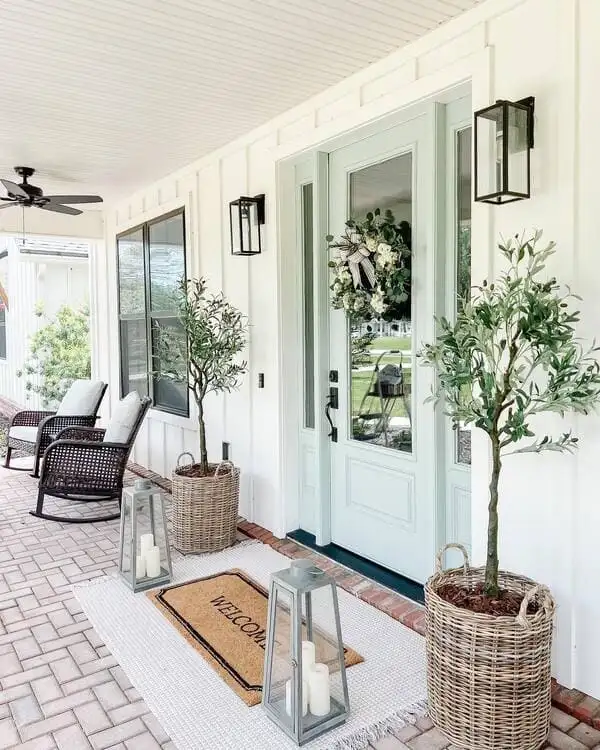 A farmhouse-style porch with board-and-batten siding, mint green front doors, wicker planters holding olive trees and oversized candle lanterns.