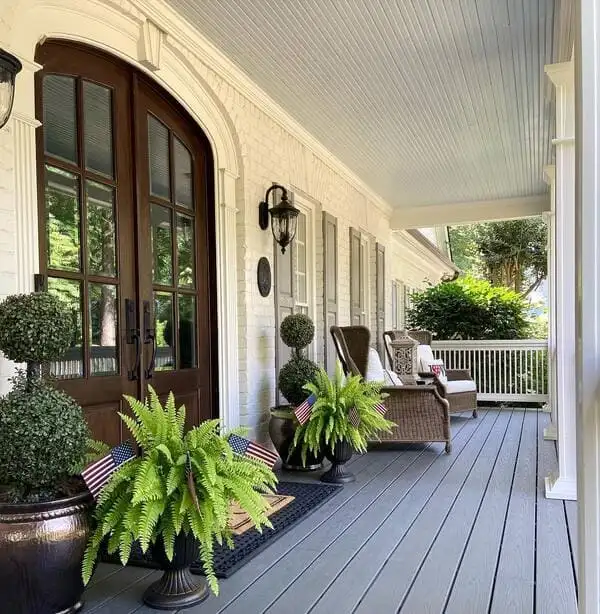 A Southern-style porch with pale blue ceiling, double dark doors, wicker chairs, ferns, and topiary planters.