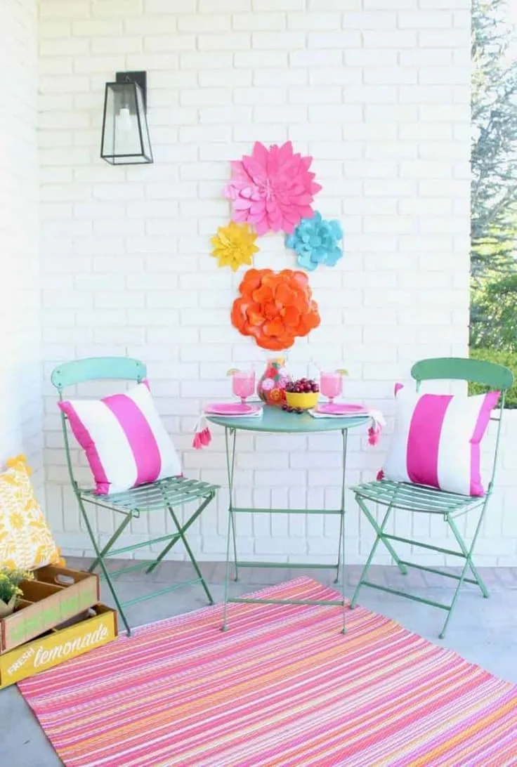 A white brick patio with bright pink striped pillows, a striped rug, and colorful paper flowers on the wall.