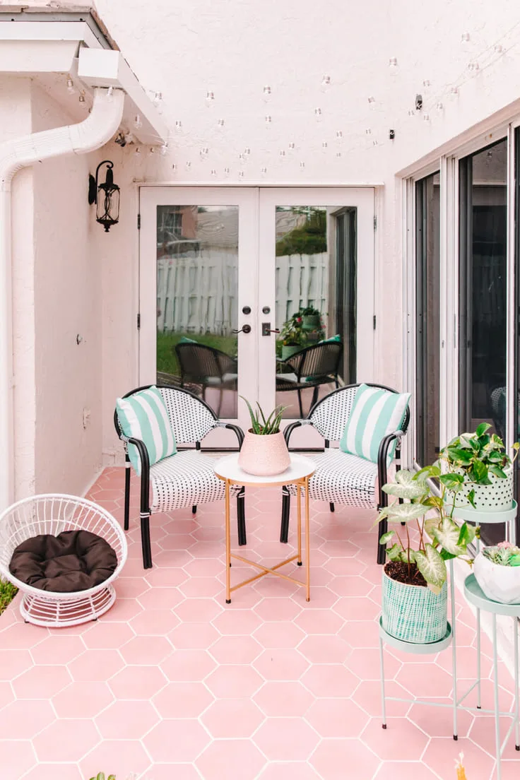A cozy outdoor patio with pink hexagonal tiles, mint green chairs with striped pillows, a small white and gold table, and potted greenery.