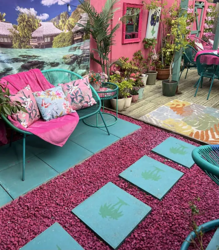A lively pink patio featuring bright green and pink chairs, tropical print pillows, potted plants, pink pebbles, and a vibrant mural backdrop.