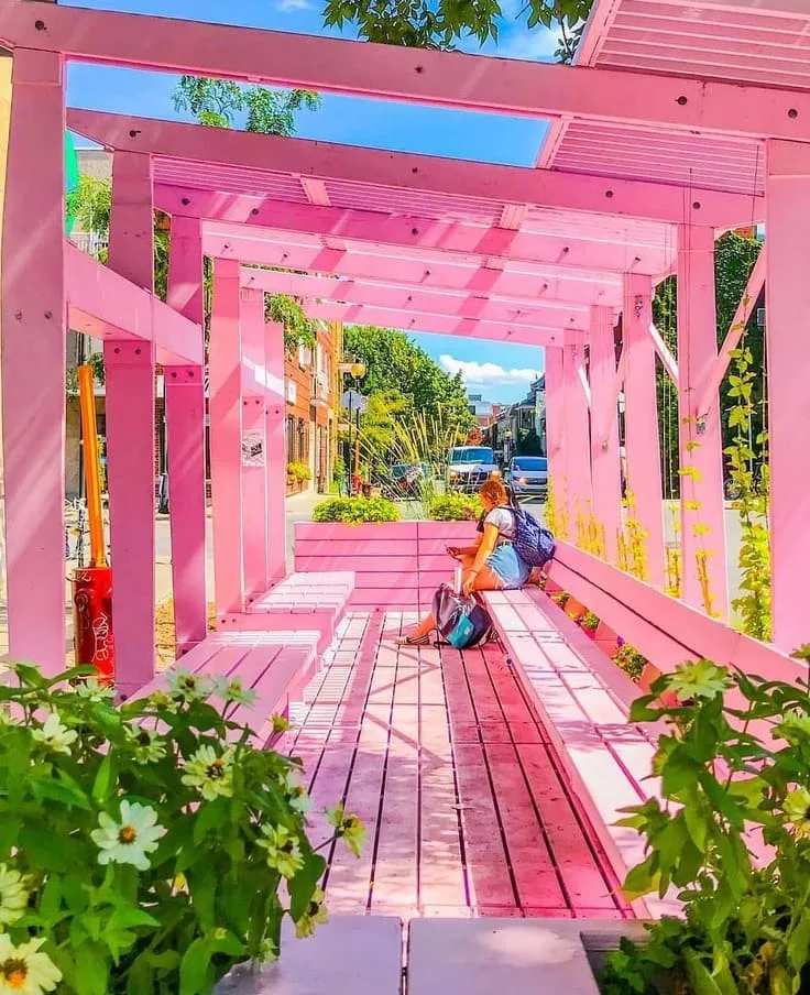 A vivid pink pergola with wooden beams and benches, providing a shaded spot in an urban street setting.