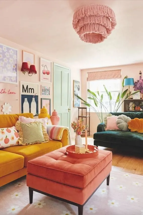 A colorful and eclectic living room featuring pastel pink walls, a pink fringed ceiling light, mustard yellow and green sofas, and a coral ottoman. Decor includes playful wall art and bold accessories.
